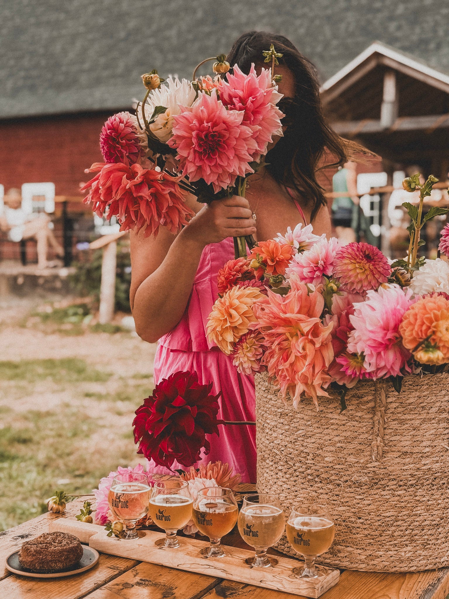 Woman in a pink dress holding flowers next to a table with drinks and food outdoors.